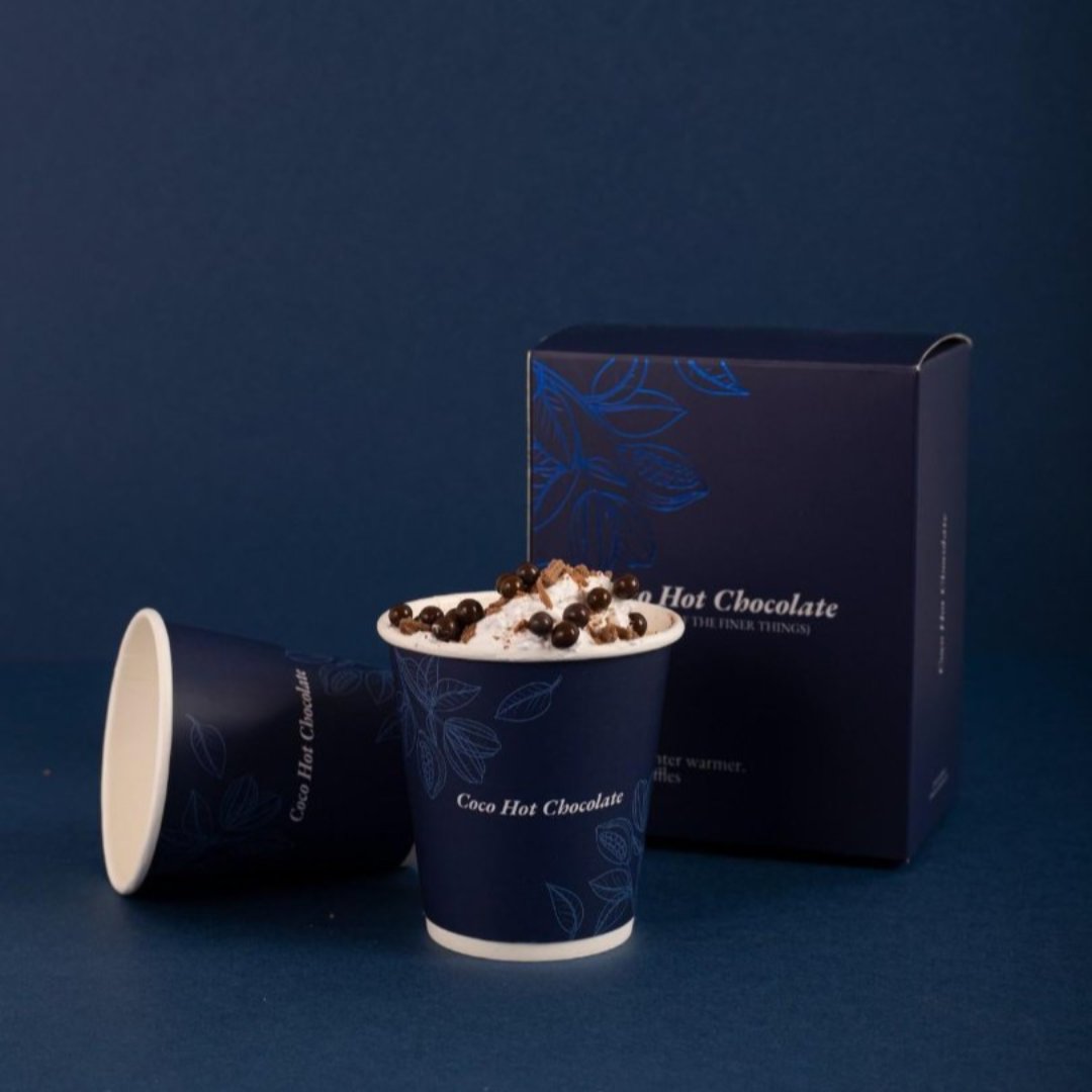 A cup of Coco Hot Chocolate with marshmallows, next to its blue packaging box with floral design and the product name visible.